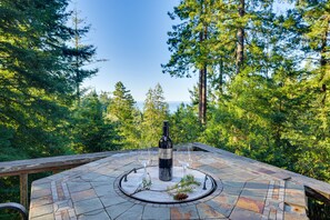 A stone-tiled outdoor seating area with a bottle of wine and glasses, surrounded by a forest.