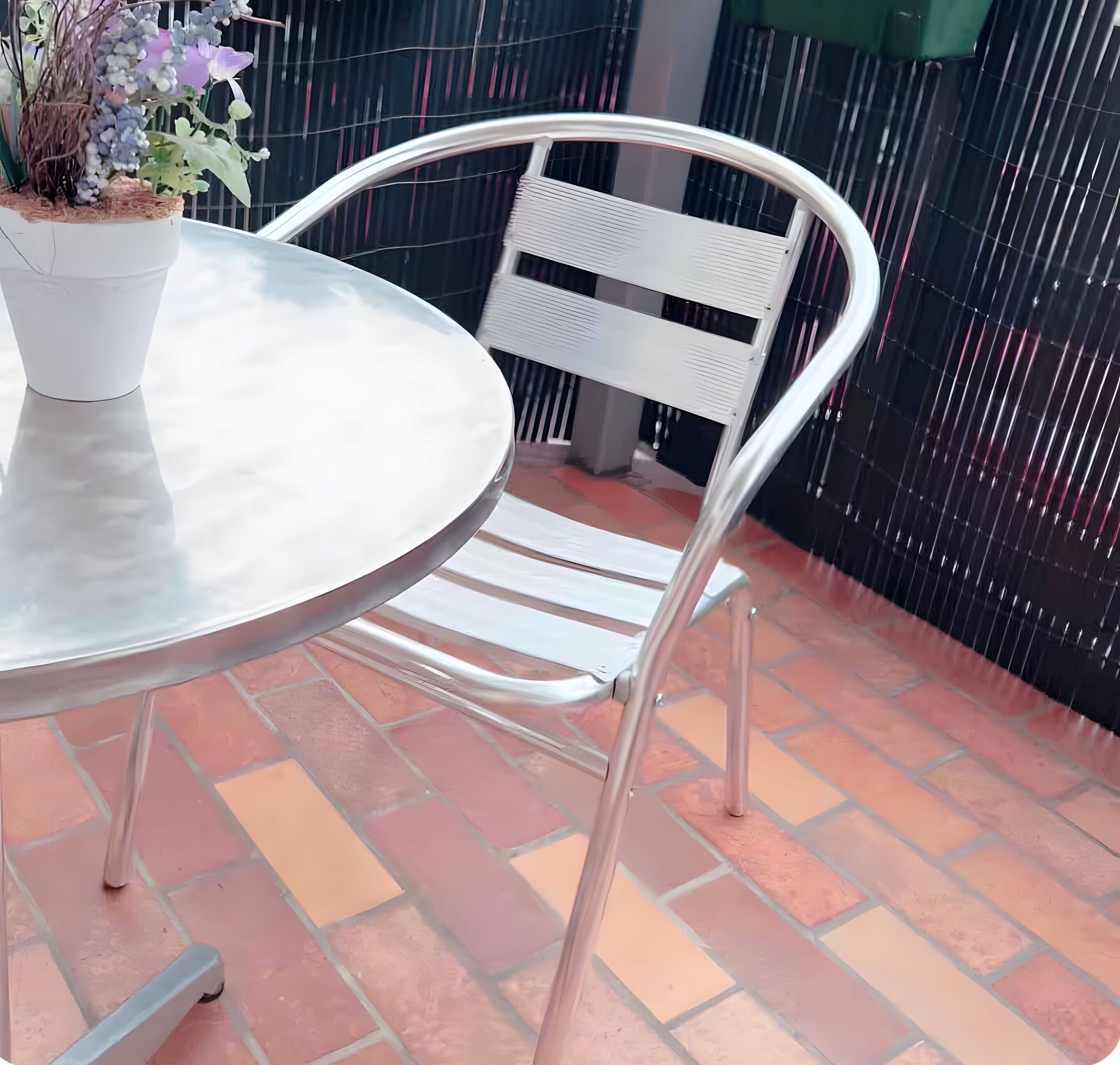 Ein runder Metalltisch mit einem Blumentopf daneben, ein weißer Metallstuhl und eine Pflasterterrasse.