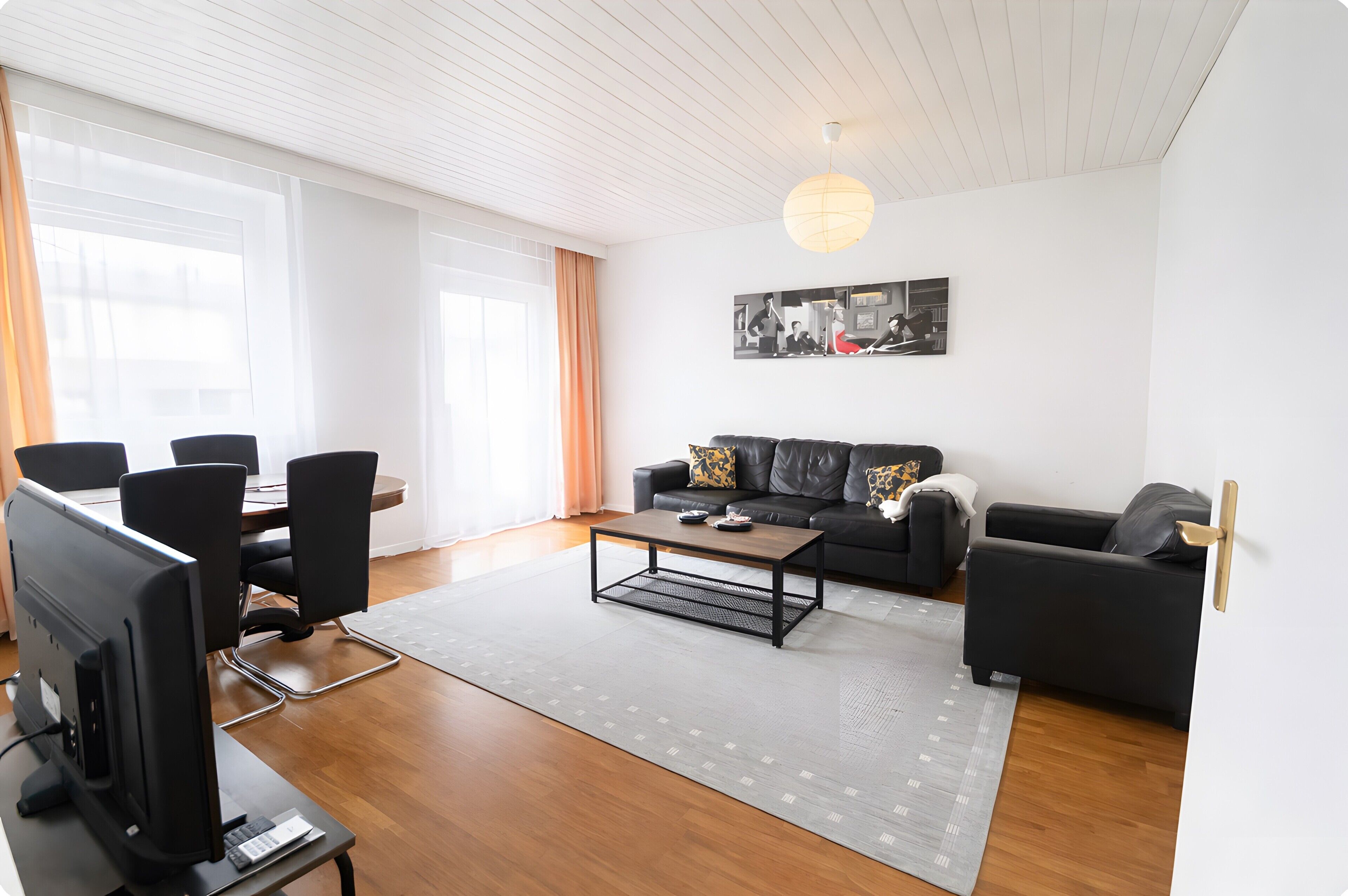 Ein modernes Wohnzimmer mit einer schwarzen Ledersofa, einem Holztisch, einer Essecke mit Tisch und Stühlen sowie einem großen Fenster mit Vorhängen.