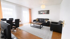 A modern living room with a black leather sofa, a wooden coffee table, a dining area with a table and chairs, and a large window with curtains. - Apartment 'Dream Home' with Balcony and Wi-Fi (Friedrichshafen)