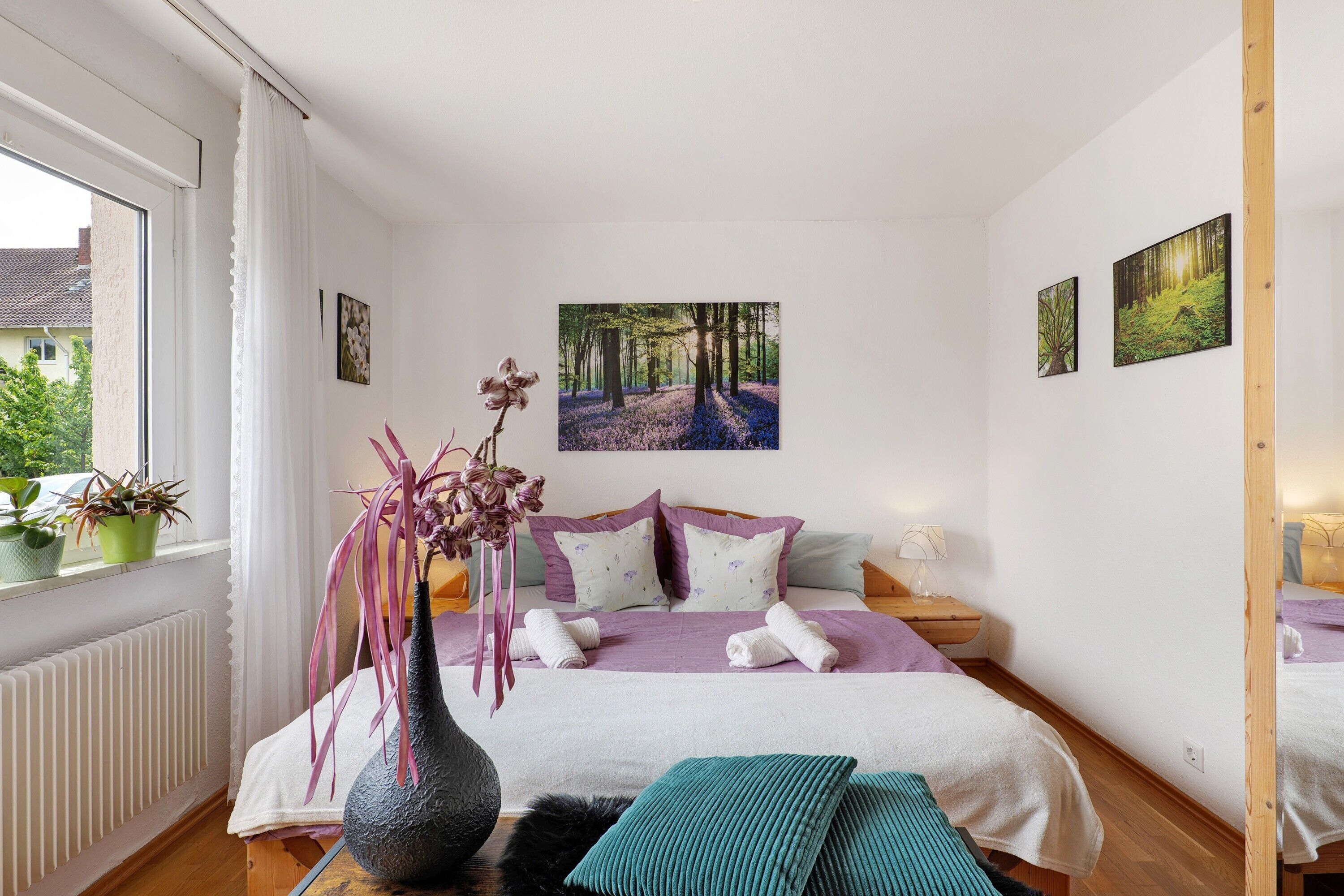Ein Schlafzimmer mit Bett, einem fensterbeprägten Fenster, einer Blumenvase und zwei Bildern an der Wand.