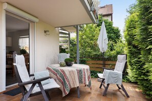 A patio with a table, two chairs, a potted plant, and a covered area.