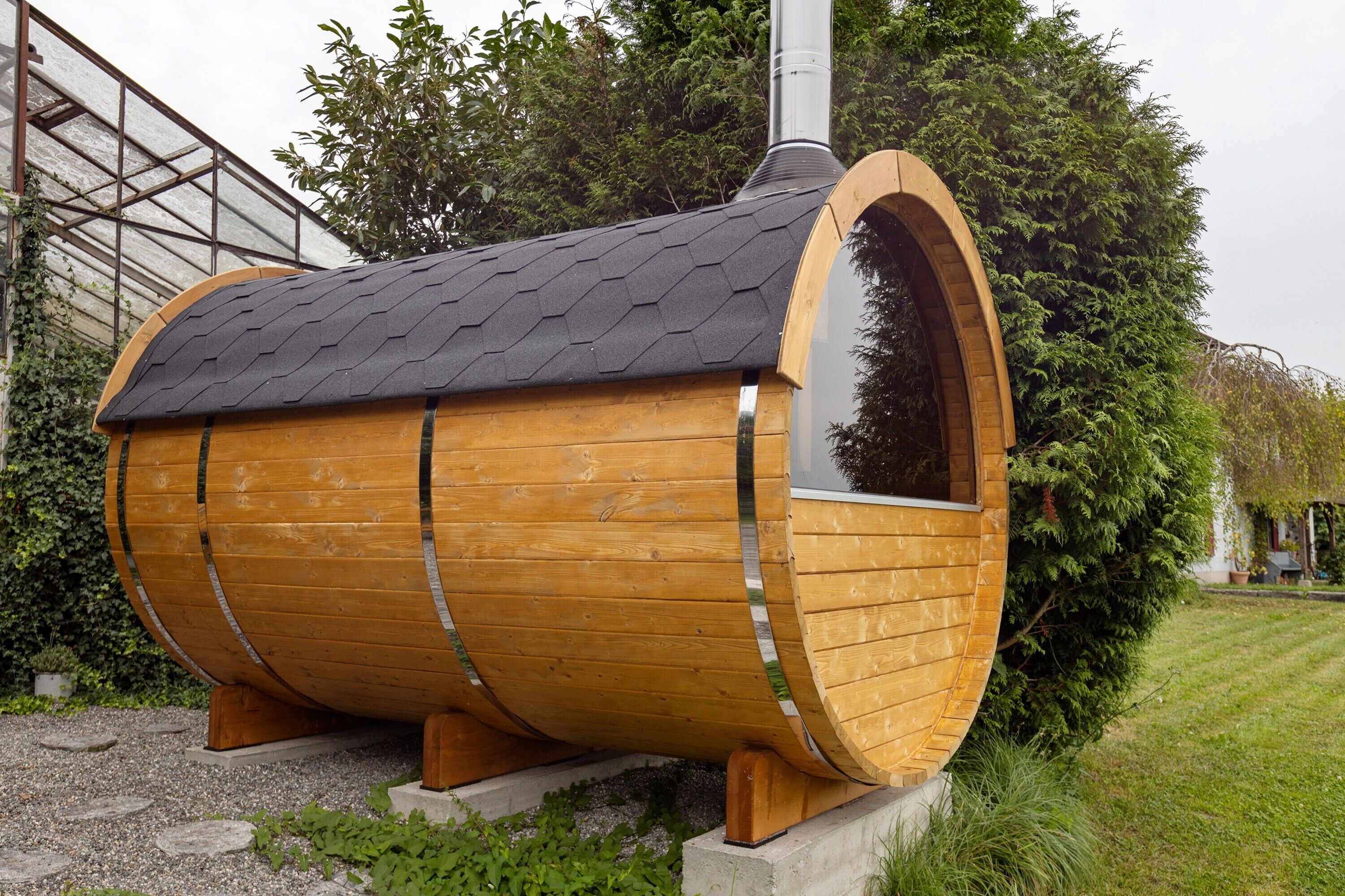 A wooden barrel-shaped structure with a chimney, surrounded by greenery and a gravel path.