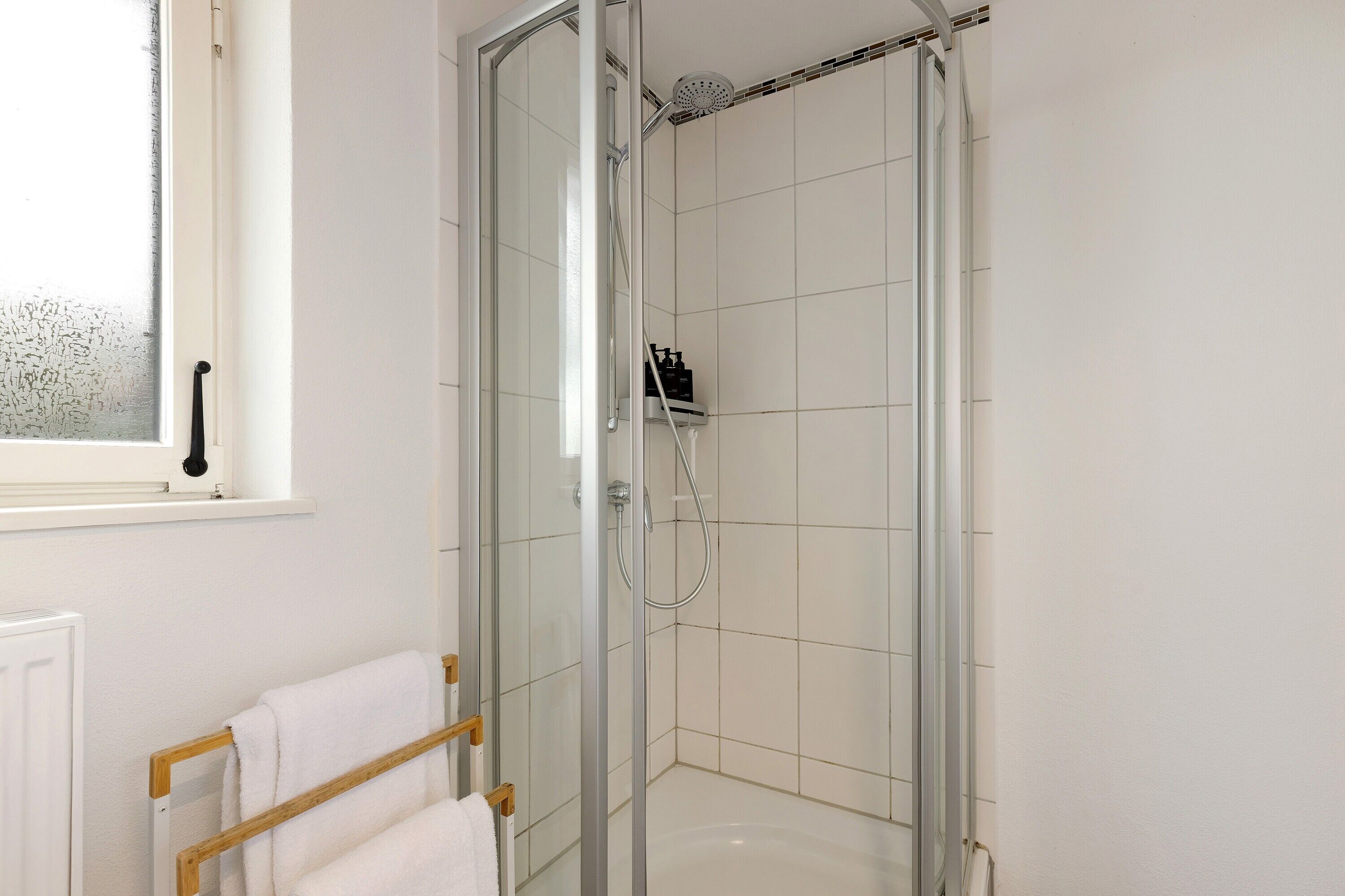 A glass-enclosed shower room with a white tiled wall, a window with frosted glass, and a towel rack with white towels.