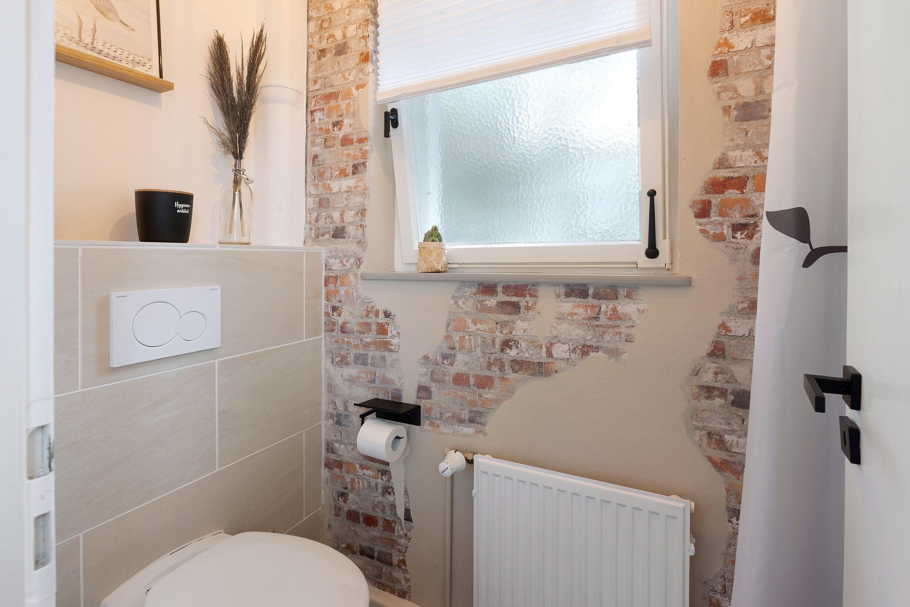 Ein Badezimmer mit Backsteinwand, Fenster, Toilette, Heizkörper und Ablagefläche mit Pflanze.
