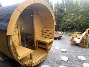 A wooden sauna with a bench and a chair, surrounded by a gravel area and trees.