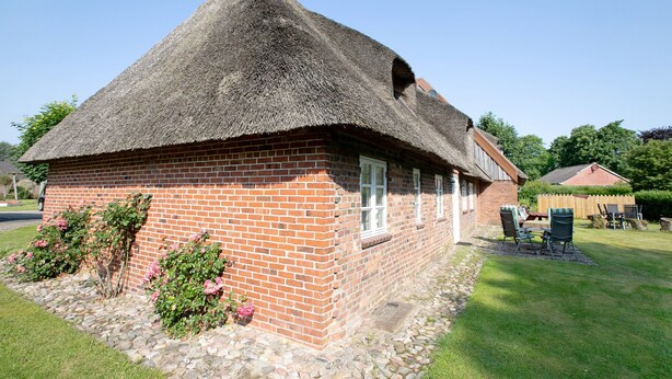 Een huis van baksteen met rieten dak, een stenen fundering en een kleine tuin met roze bloemen.