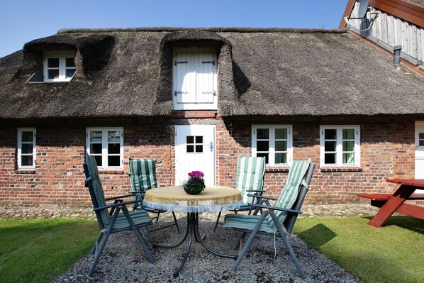 A thatched-roof cottage with a brick exterior, a round table with chairs, and a wooden picnic table.