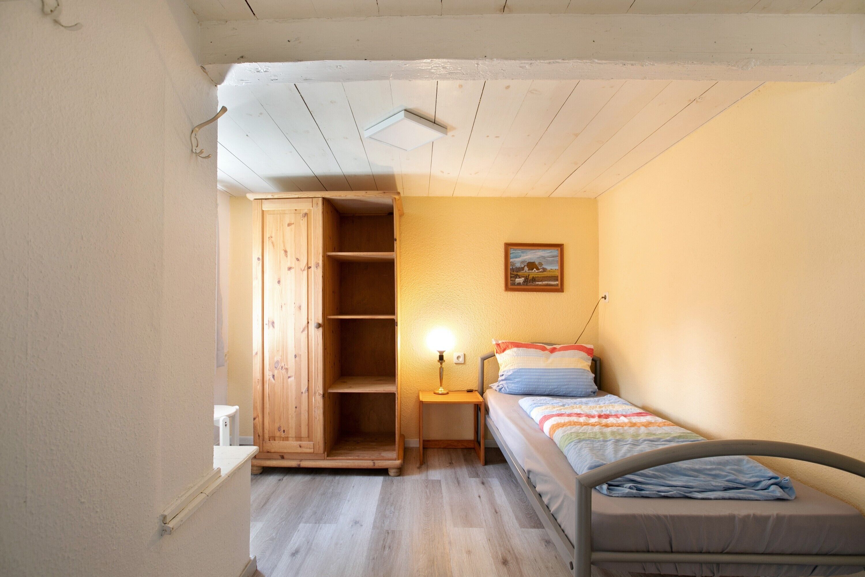 A bedroom with a bed, a wooden wardrobe, a nightstand with a lamp, and a painting on the wall.