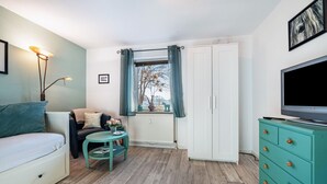 Una habitación con sofá, una mesita con un florero, un mueble con cajones, televisor y ventana con cortinas.