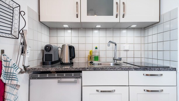 Una cocina con armarios blancos, encimera de granito, cafetera, hervidor, lavamanos y escurreplatos.