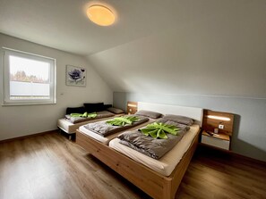 Une chambre avec un plafond en pente, une fenêtre avec des stores, un lit avec des oreillers et une couverture, une table de chevet avec une lampe, et une œuvre d’art accrochée au mur.