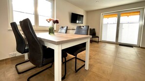 Ein Essbereich mit Tisch, Stühlen, einer Vase mit Blumen, einem Fernseher und großen Fenstern.