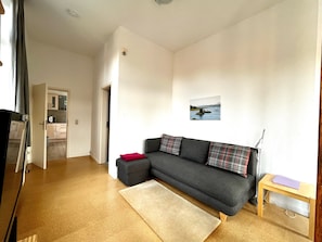 Ein Wohnzimmer mit einer Couch, einem kleinen Tisch, einem Teppich und einer durchgehenden Tür erkennbaren Küche.