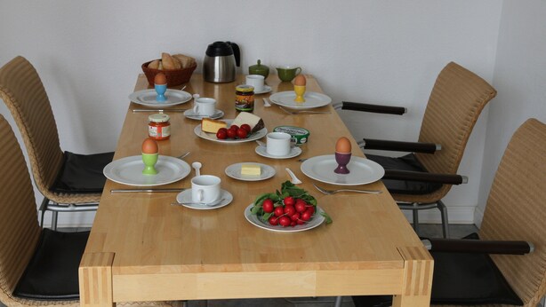 Een eettafel gedekt met borden, kopjes en diverse soorten eten, omringd door rieten stoelen.