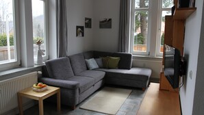 Een moderne woonkamer met een grijze hoekbank, een klein houten tafeltje met fruit, en een televisie op een dressoir.