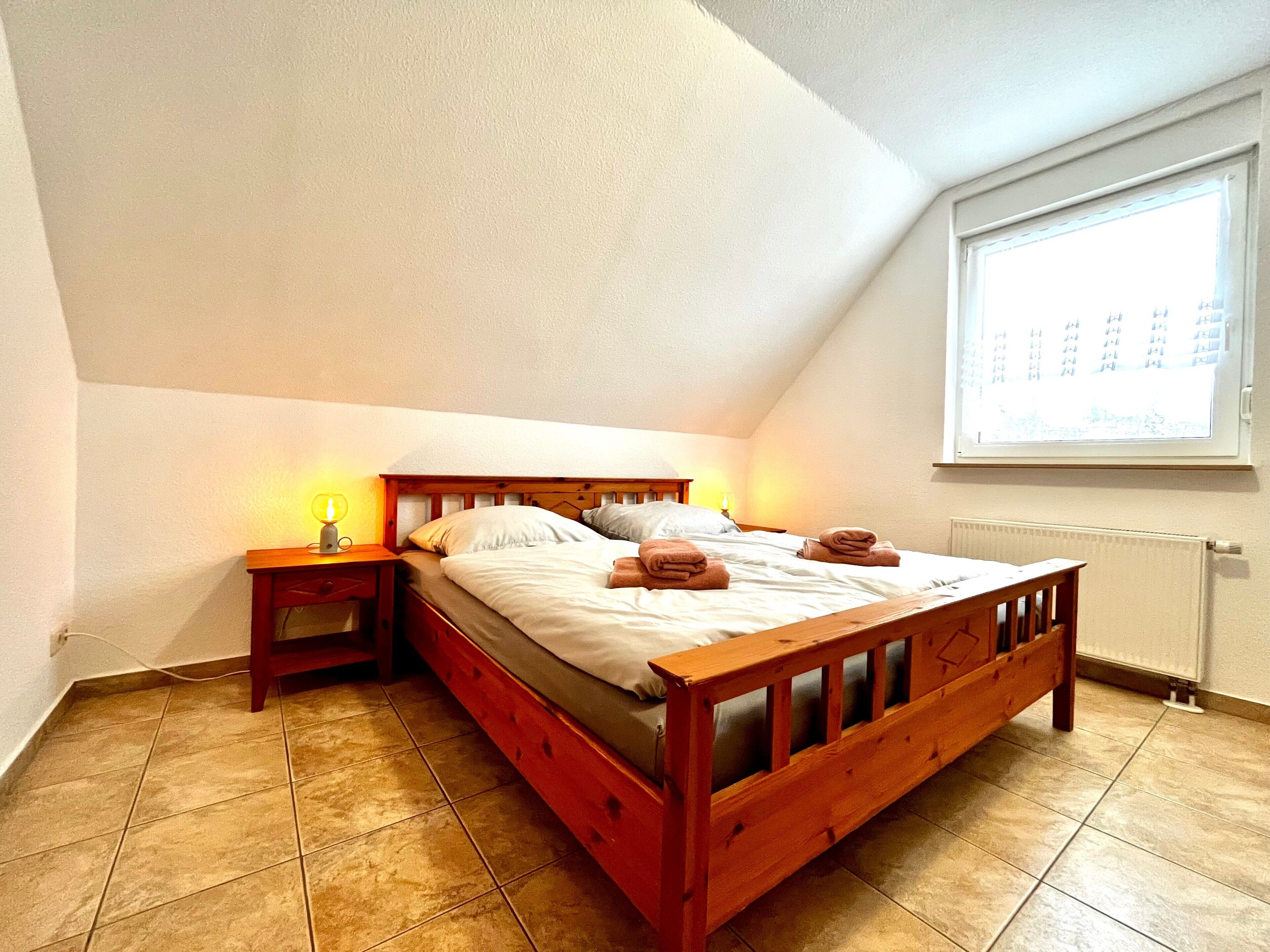 A bedroom with a wooden bed, two bedside tables with lamps, a window with blinds, and a radiator.