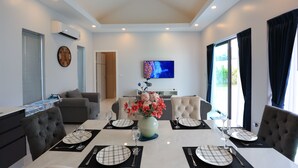 Een moderne eetruimte met een witte tafel gedekt voor vier personen, een bloevas en een flatscreen televisie aan de muur.