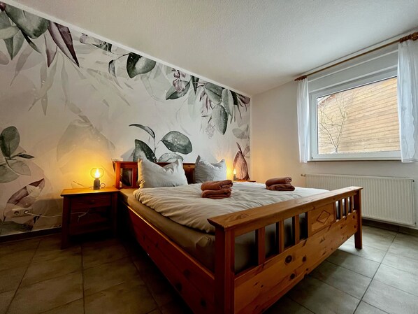 Un dormitorio con cama, mesitas de noche, una ventana con persianas y una pared con diseño de hojas.