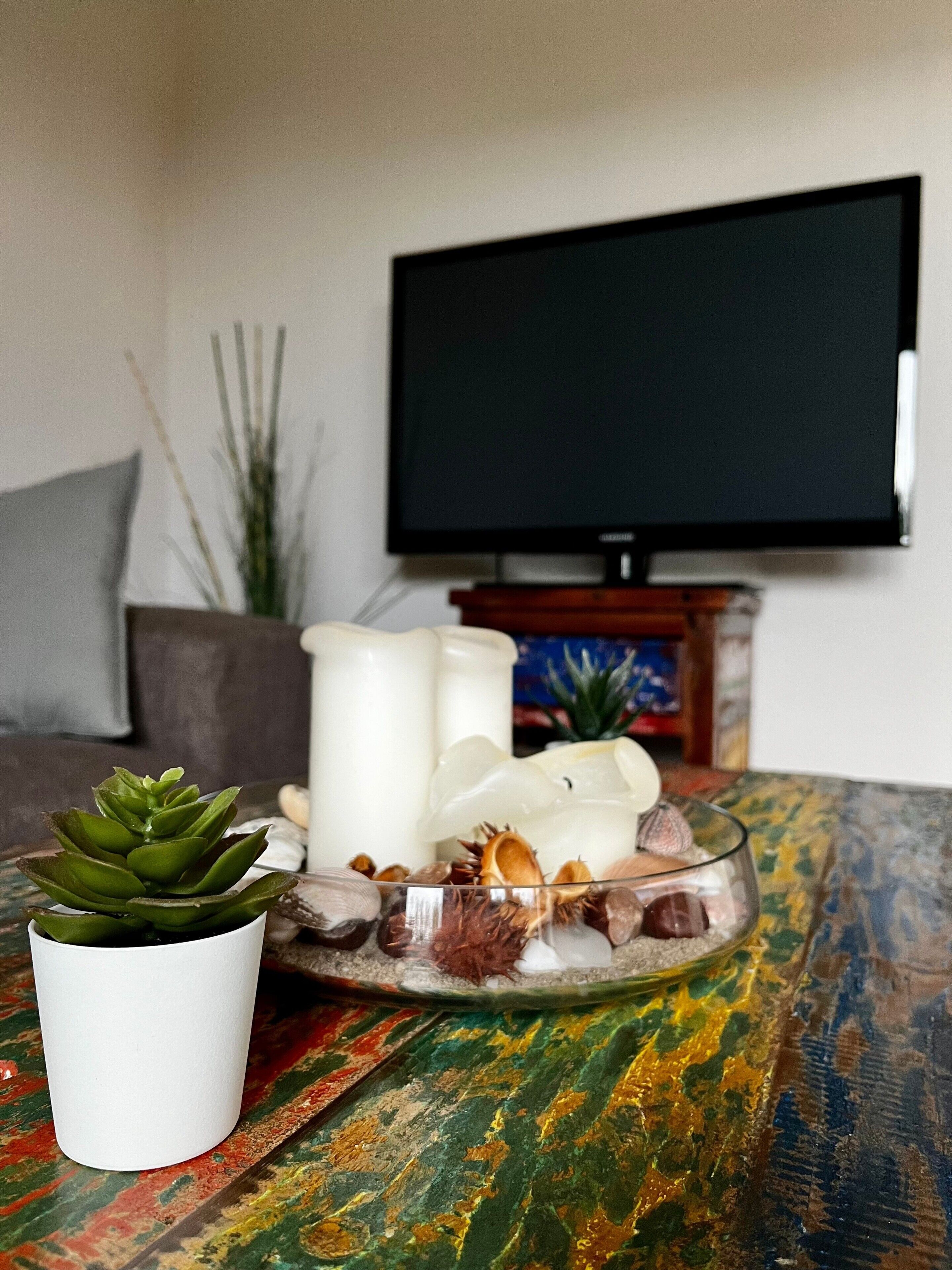 Ett vardagsrum med en platt-TV, en krukväxt av typen suckulent, ljus, snäckskal och ett färgglatt bordsduk.