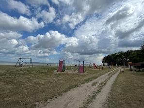 An outdoor playground with swings and climbing structures, a dirt path, and a body of water in the background. - Holiday Home 'Haus Strandräuber Mit Sauna' with Private Terrace, Private Garden and Wi-Fi (Zierow)