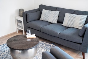 Un canapé gris agrémenté de coussins à rayures, une petite table d’appoint blanche, une table basse ronde en métal et un tapis à motifs.