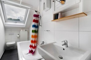 A modern bathroom with a skylight, a shower with a colorful curtain, a white sink, and a wooden shelf.