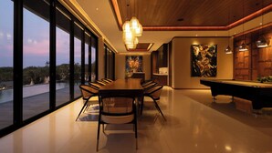Un espace repas moderne avec une longue table et des chaises, un billard et de grandes fenêtres donnant sur l’extérieur.