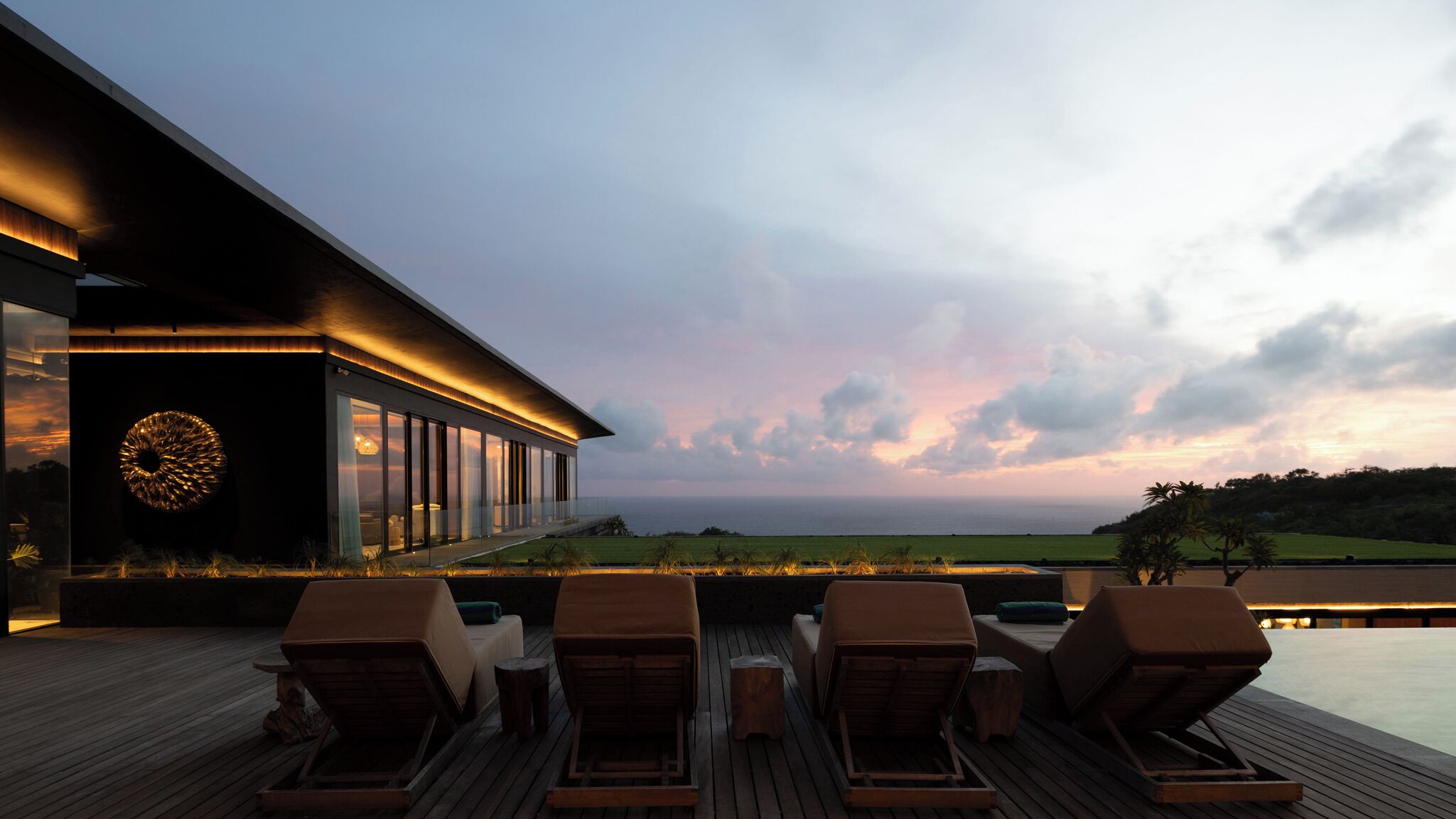 A modern building with a large glass facade, a deck with lounge chairs, and a view of the ocean at sunset.