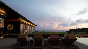 Un bâtiment moderne doté d’une grande façade vitrée, d’une terrasse avec des chaises longues et offrant une vue sur l’océan au coucher du soleil.