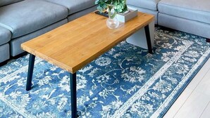 Ein modernes Wohnzimmer mit einem grauen Ecksofa, einem Holztisch und einem blauen Teppich mit Muster.