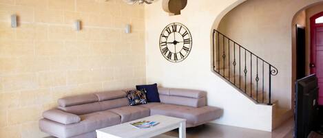 A living room with a beige sofa, a white coffee table, a wall clock, a chandelier, and a staircase with a red door.