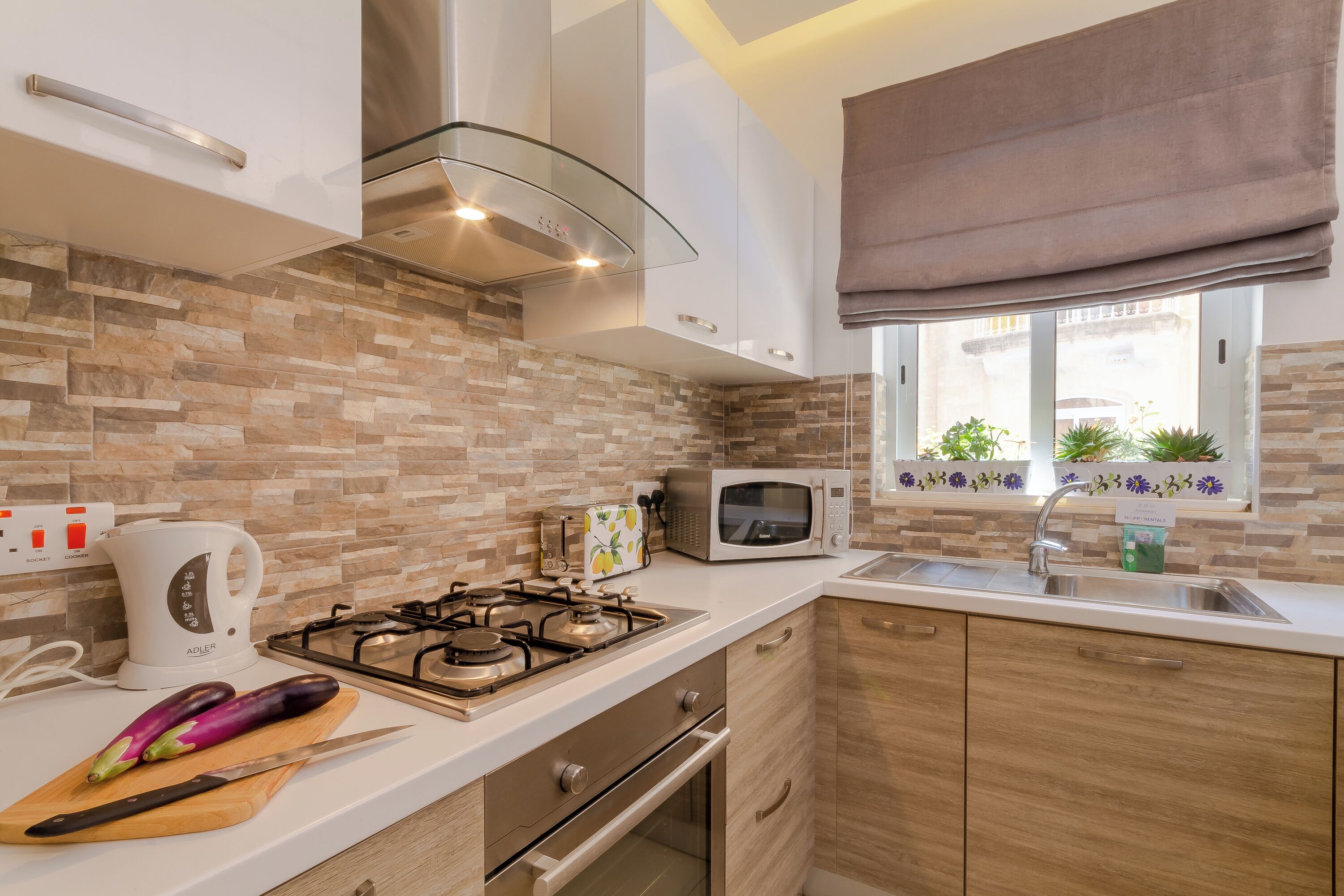 Cocina moderna con estufa a gas, microondas y tetera. Se ve una ventana con plantas en el alféizar.