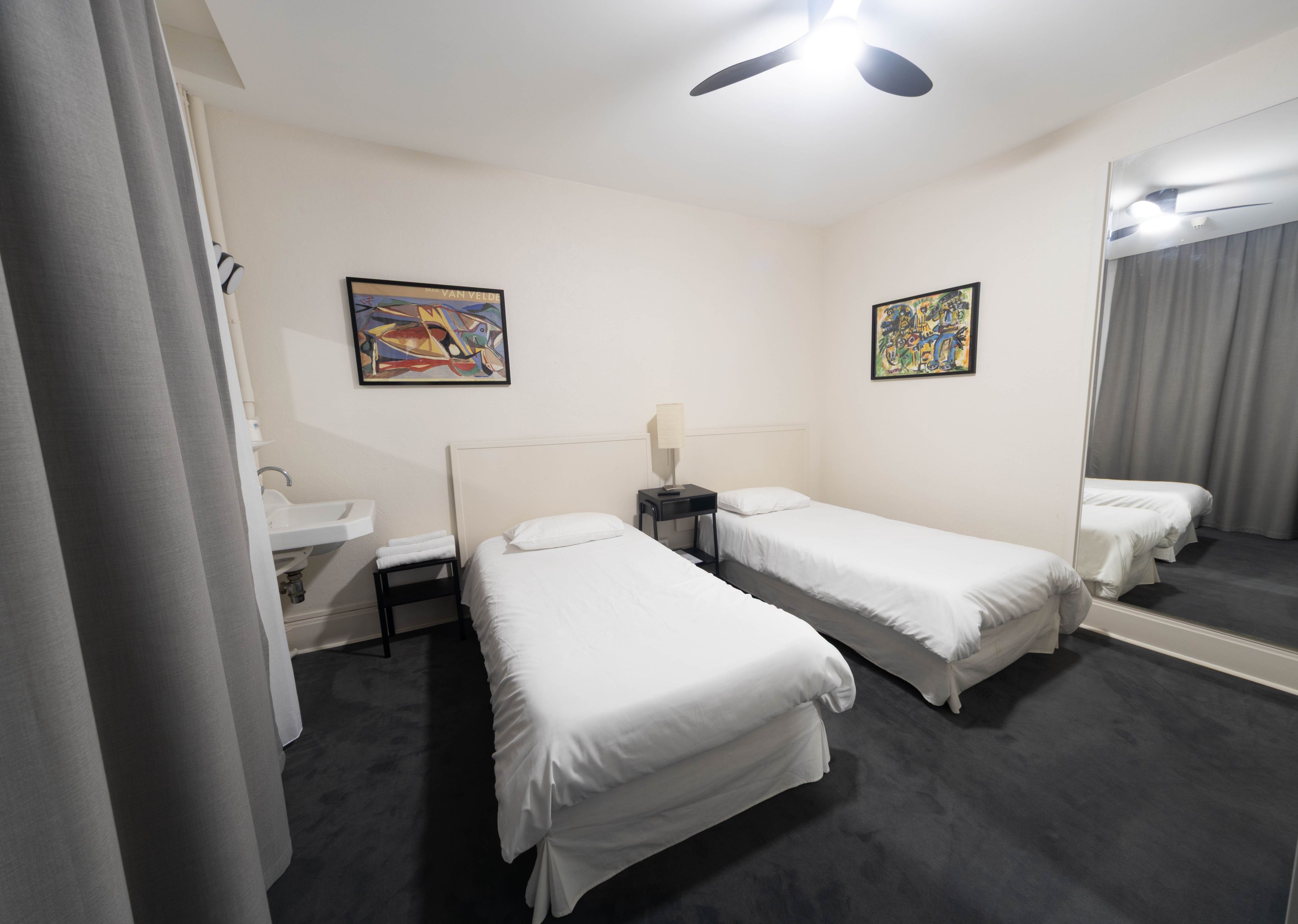 Habitación de hotel con dos camas individuales, una mesita de noche con lámpara, un espejo, un lavabo y dos cuadros abstractos.