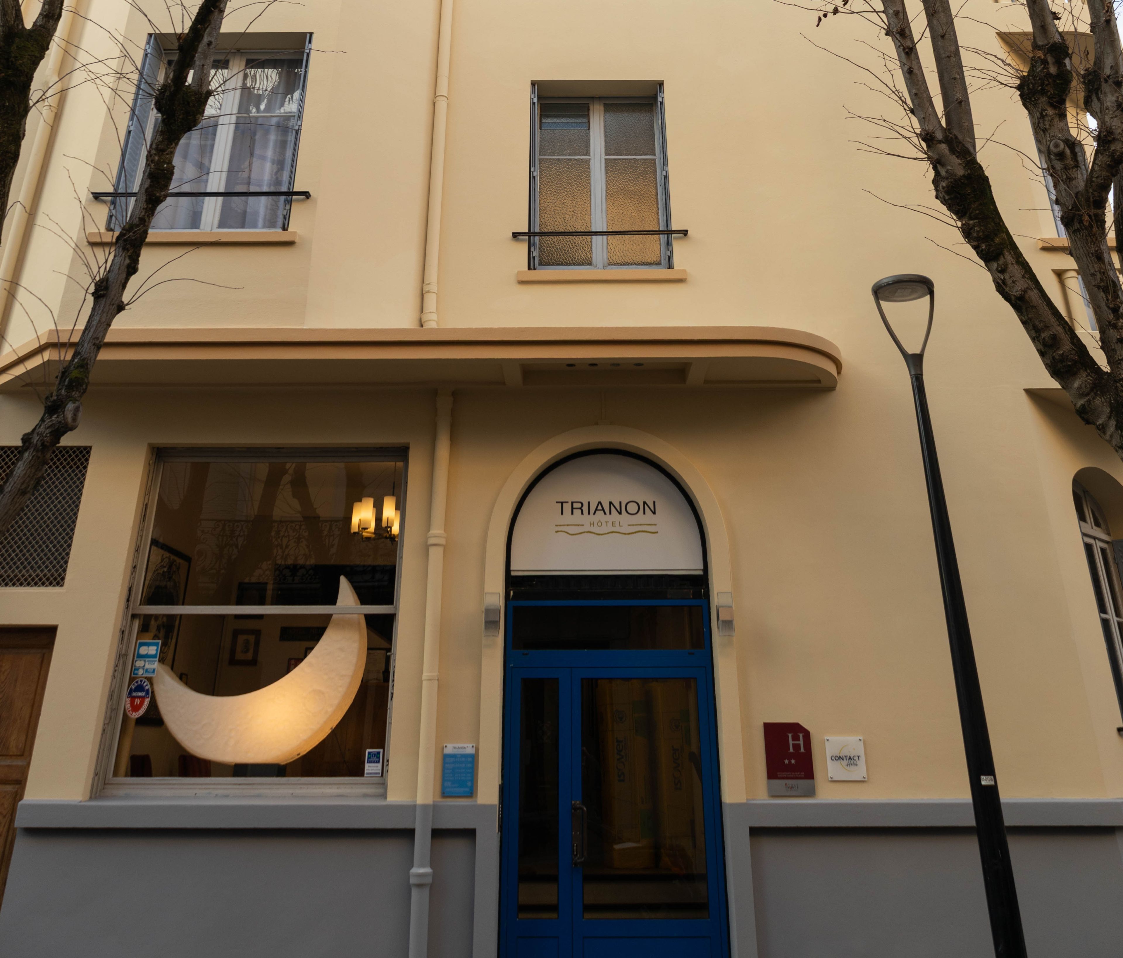 Entrada de un hotel con una puerta azul, un ventanal en forma de media luna y un cartel que dice 'TRIANON HOTEL'.