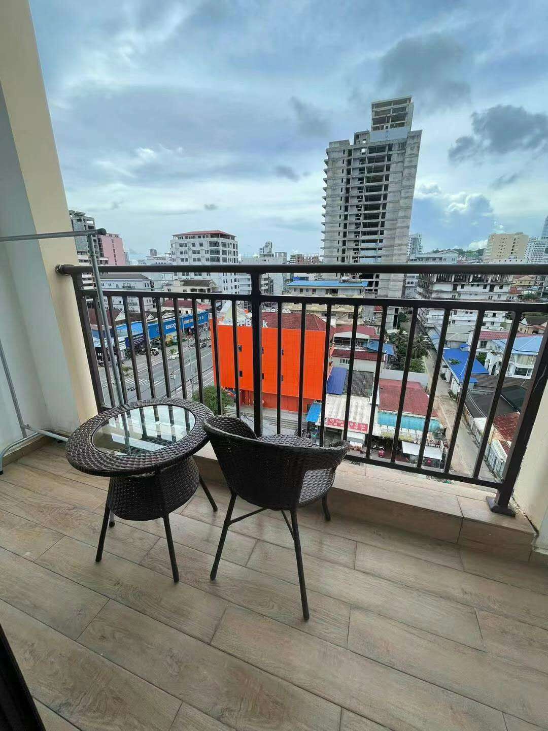 A balcony with a glass table and two chairs, overlooking a cityscape with buildings and a clear sky.