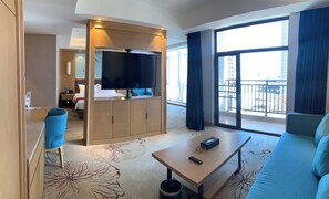 A hotel room with a large TV, a sofa, a coffee table, a desk, and a balcony with a view.