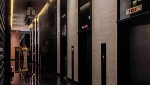 A modern hotel corridor with reflective black tiles, a central elevator, and a decorative vase in the distance.