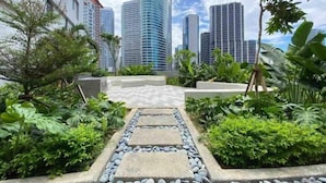A pathway leading to a modern building with a curved facade, surrounded by lush greenery and other high-rise buildings in the background.