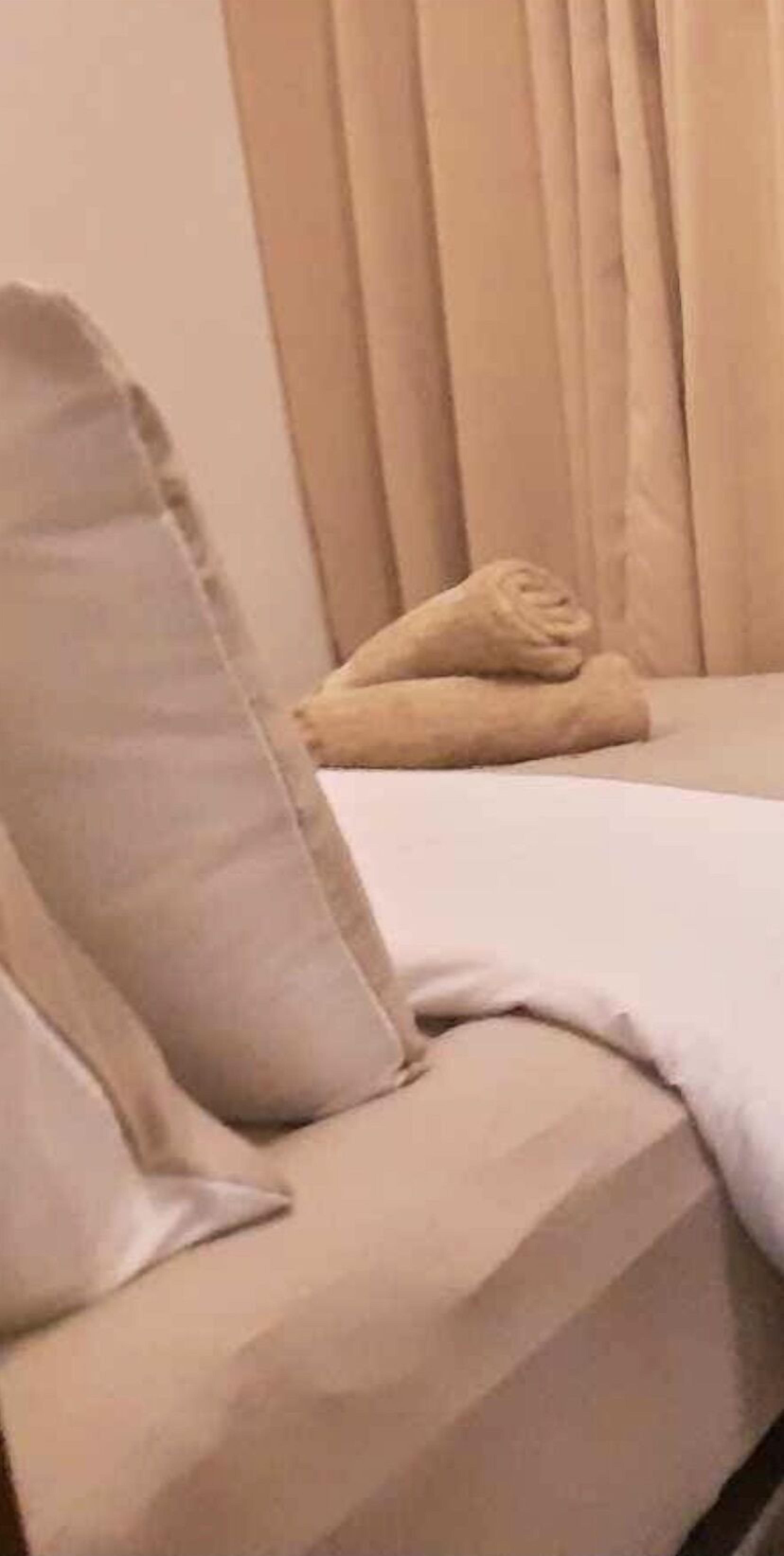 Uma cama com colchão e almofadas bege, um cobertor dobrado e uma cortina vertical ao fundo.