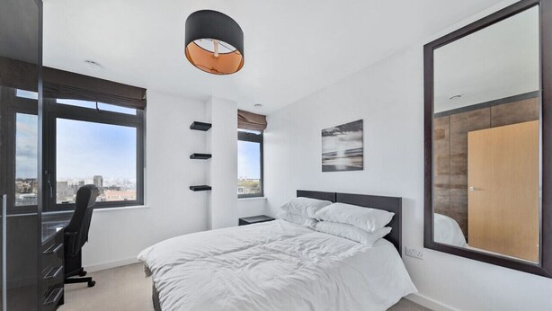 A modern bedroom with a large bed, a desk, and a chair. There are multiple windows providing a view of the city.