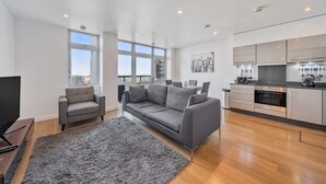 A modern living room with a grey sofa, a small armchair, and a large rug. There is a kitchen area with stainless steel appliances and a dining table with chairs.