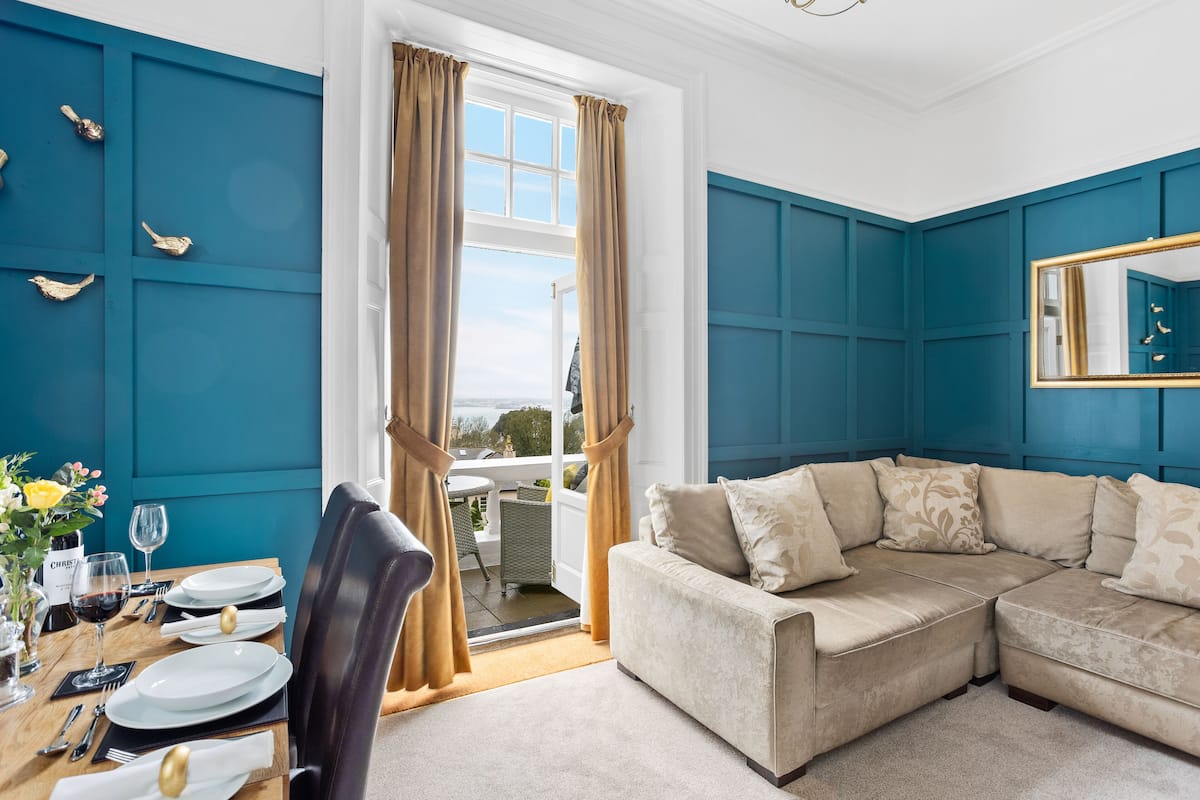 A room with teal walls, a beige sectional sofa, a dining table set with plates and glasses, and a window with curtains.