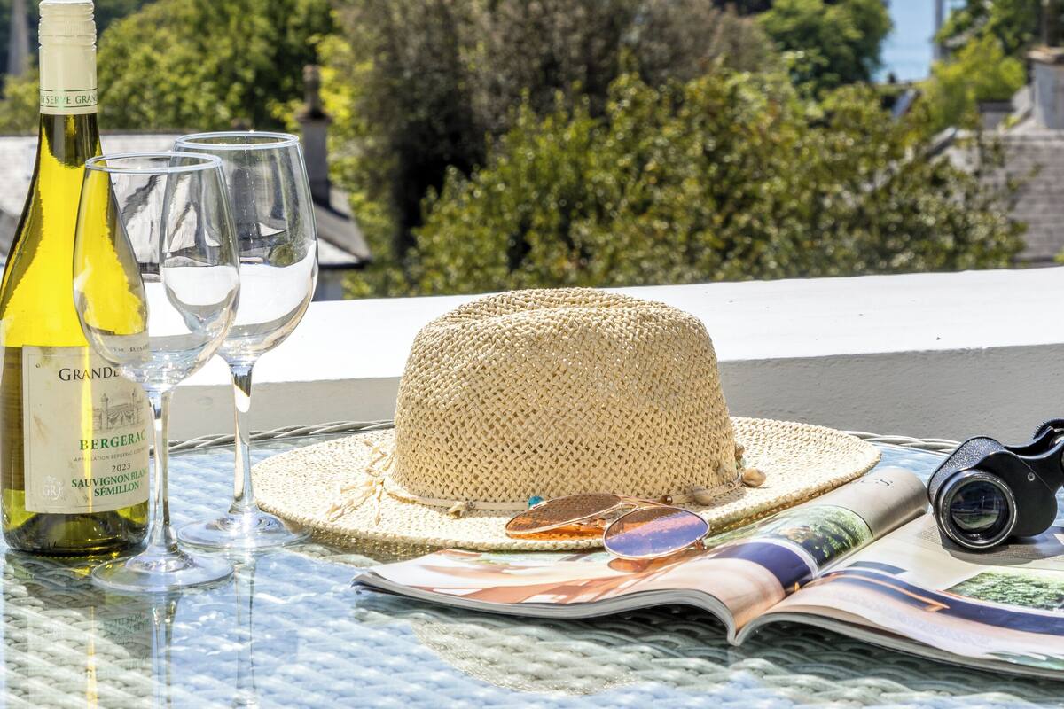 Une table avec une bouteille de vin, des verres, un chapeau de paille et un appareil photo, sur un balcon donnant sur une étendue d’eau et une zone verdoyante.