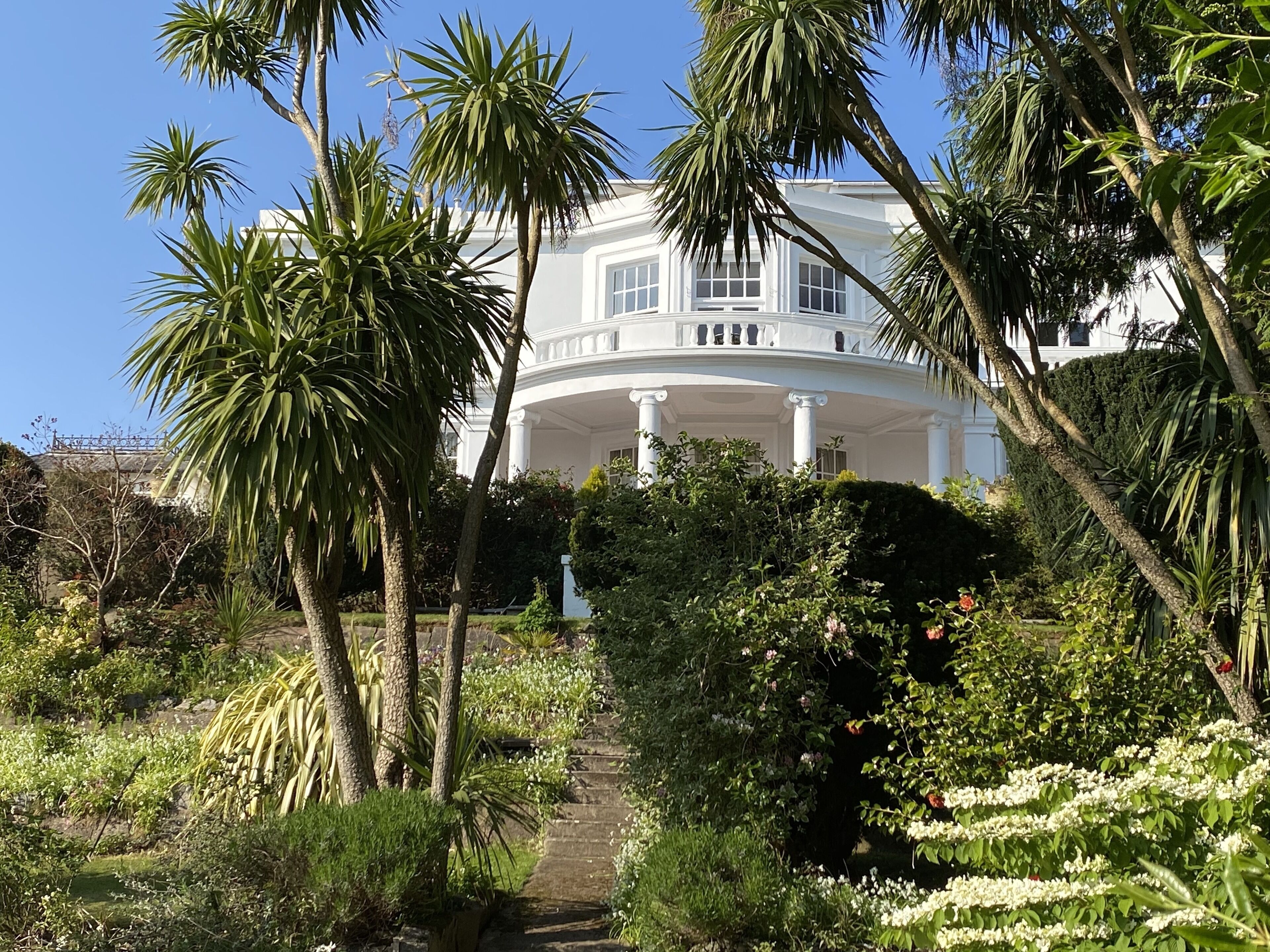 A large, white, classical-style building with a central circular porch and columns, surrounded by lush greenery and palm trees.