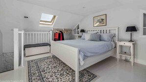 A bedroom with a white bed, a bench, a nightstand, a lamp, a wardrobe, and a skylight.