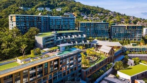 A modern building complex with multiple high-rise structures, surrounded by lush greenery and a hilly backdrop. - The Panora apartments by NLA (Choeng Thale)