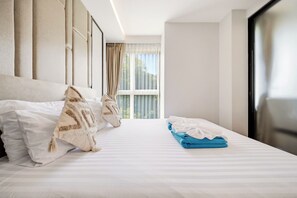 A hotel room with a large bed, two pillows, a blue towel, and a window with curtains. - The Panora apartments by NLA (Choeng Thale)