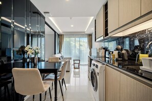 A modern kitchen with a built-in washing machine, a dining area with a table and chairs, and a large window with curtains. - The Panora apartments by NLA (Choeng Thale)