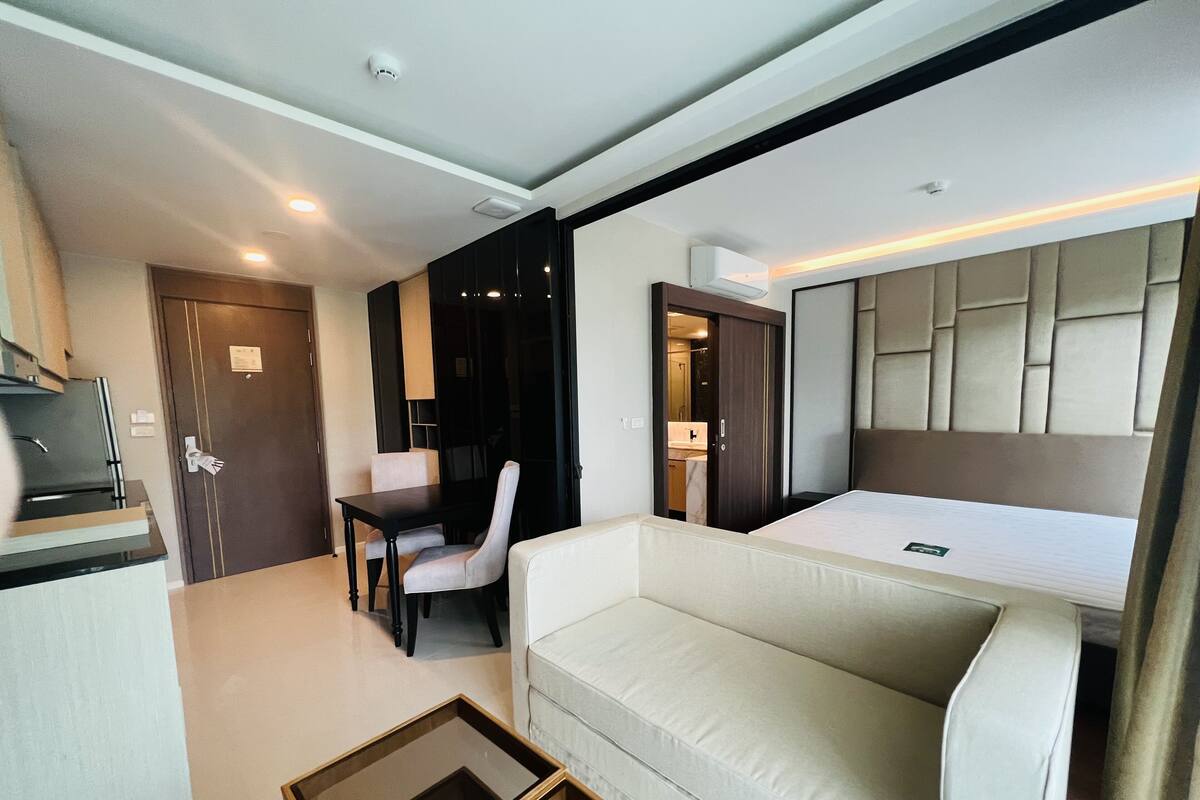 A modern hotel room with a sofa, a small table, a desk with a chair, a bed, and a kitchenette area.